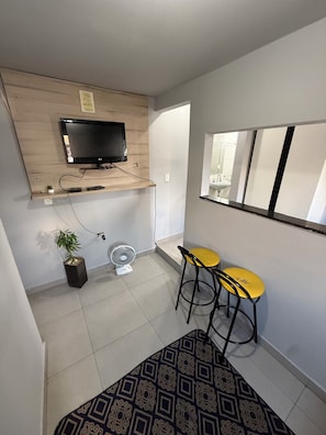 A small, modern room with a TV mounted on a wooden shelf, a small fan, two yellow stools, and a patterned rug. - Comfortable Furnished Apartment in the North Zone of São Paulo (São Paulo)