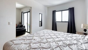 Un dormitorio con cama, ventana con cortinas, puerta que da a otra habitación y una lámpara en una mesita de noche.