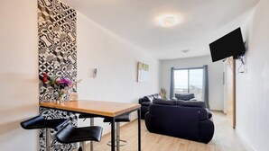 Una sala de estar moderna con un sofá, una mesa de comedor con dos sillas, un televisor montado en la pared y un ventanal grande.
