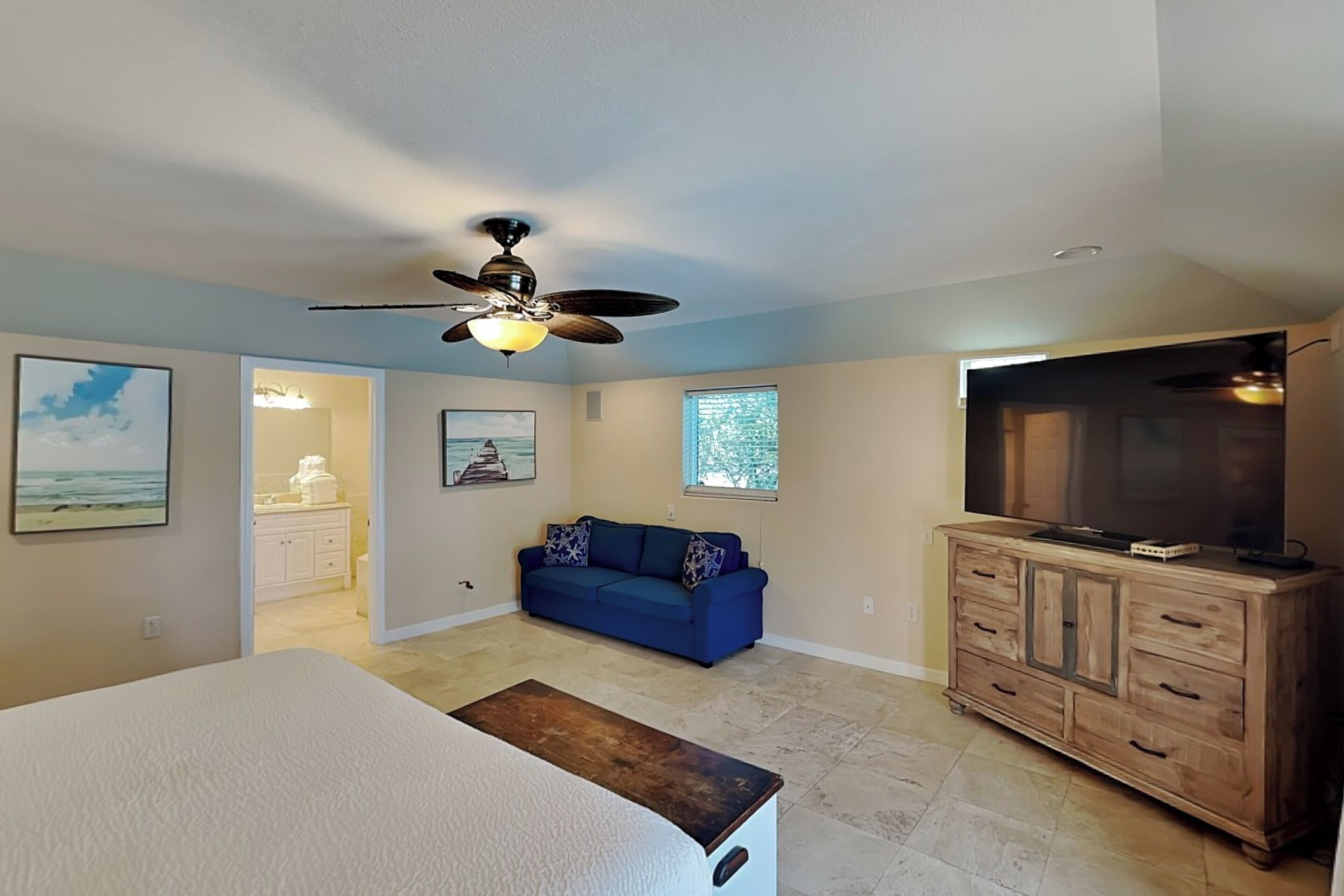 A bedroom with a bed, a ceiling fan, a dresser, a TV, a blue couch, and a window.