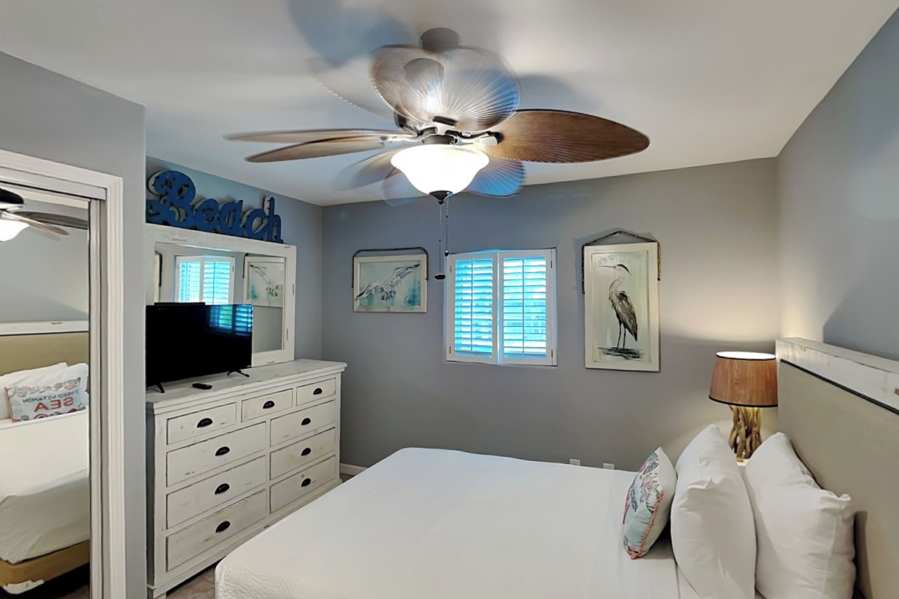 A bedroom with a bed, a dresser, a ceiling fan, and artwork on the walls.
