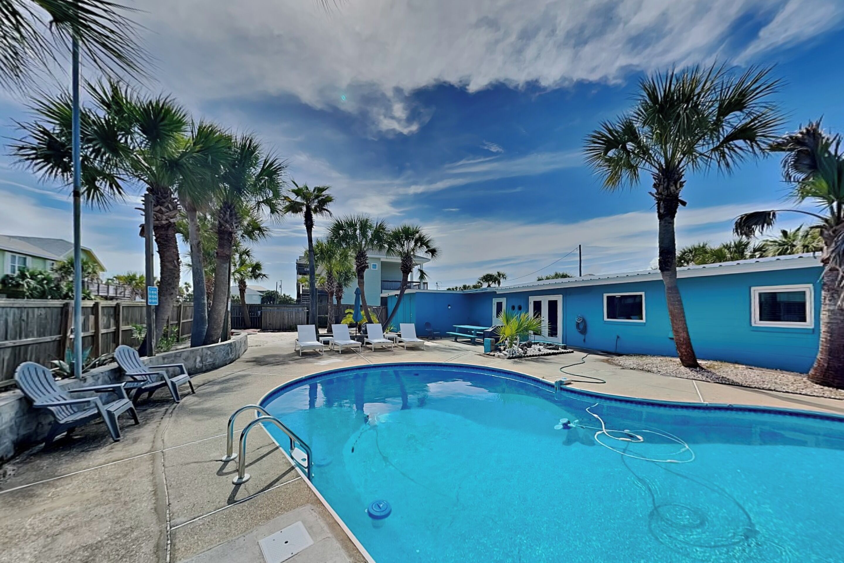 Una piscina con sillones de descanso, palmeras y un edificio azul al fondo.