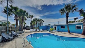 A swimming pool with lounge chairs, palm trees, and a blue building in the background. - Stay Awhile (Pensacola Beach)