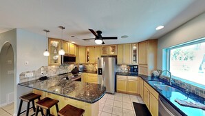 A modern kitchen with a central island, bar stools, a refrigerator, and a window with a view. - Stay Awhile (Pensacola Beach)