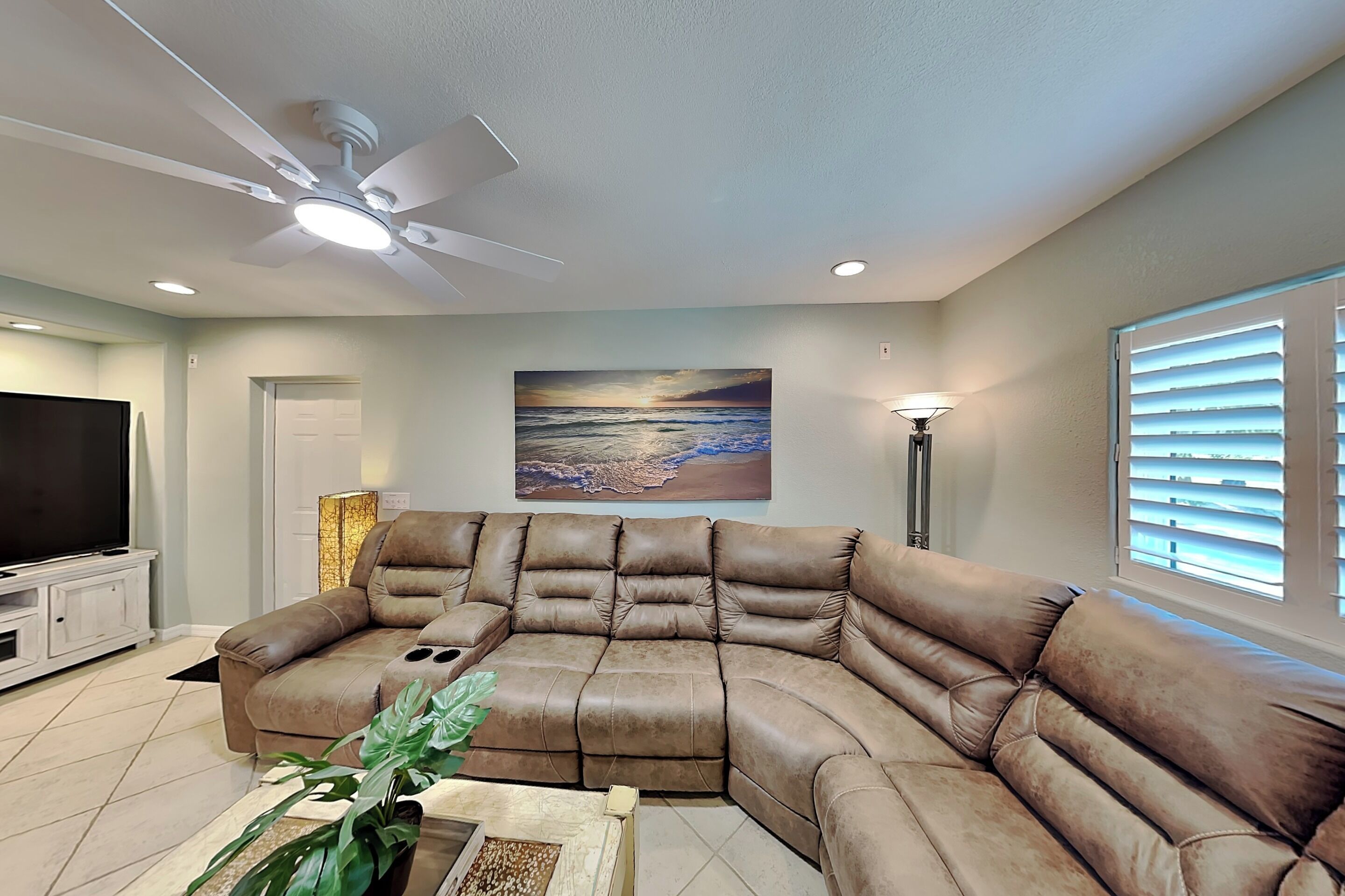A living room with a sectional sofa, ceiling fan, TV, and a painting of a beach scene.