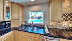 A kitchen with wooden cabinets, a double sink, a coffee maker, and a view of a swimming pool. - Stay Awhile (Pensacola Beach)