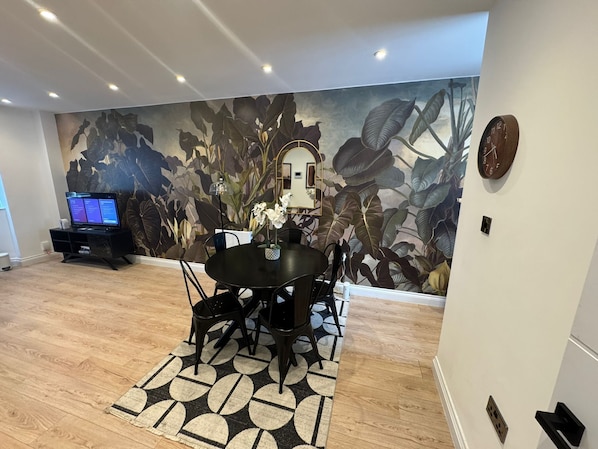 Un comedor con una mesa redonda, sillas, un mural en la pared, un televisor y un reloj.