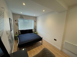 Um quarto com cama, escrivaninha, cadeira, janela com persianas e luminária na parede.
