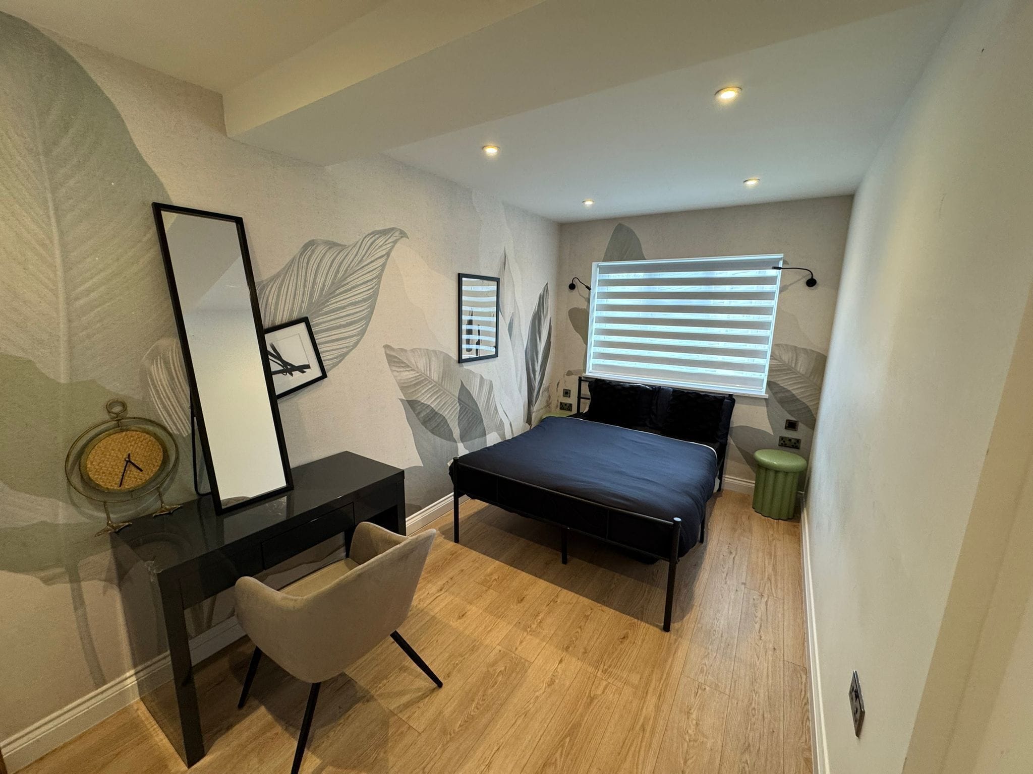 A bedroom with a bed, a desk, a chair, a window with blinds, and a wall clock.
