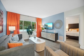 Ein modernes Wohnzimmer mit einer Couch, Sesseln, einem Couchtisch, einem Fernseher und einem Bett im Hintergrund.