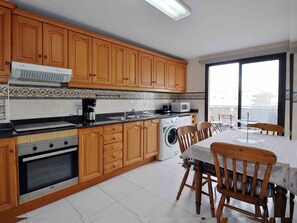A kitchen with wooden cabinets, a washing machine, a microwave, and a dining area with a table and chairs. - Roger de Flor in Puerto Pollensa (Pollença)