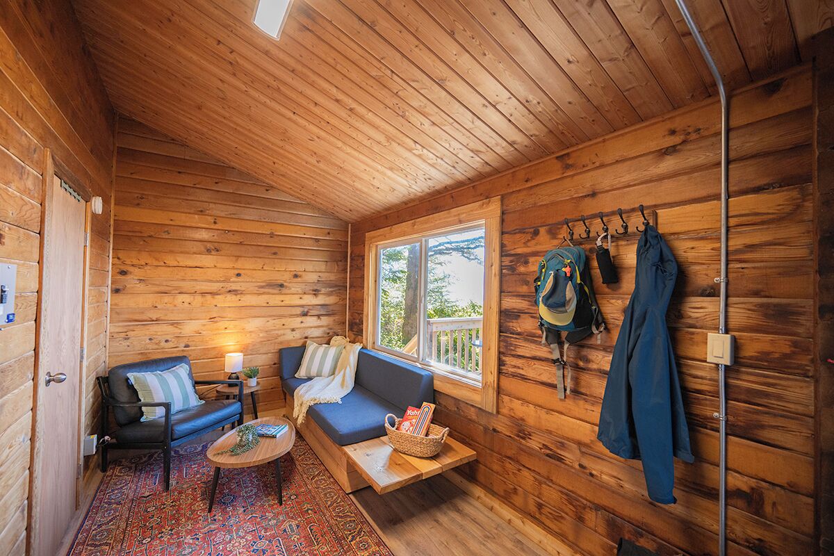 A cozy cabin interior with a sofa, a small table, a window with a view, and a coat rack with hanging items.