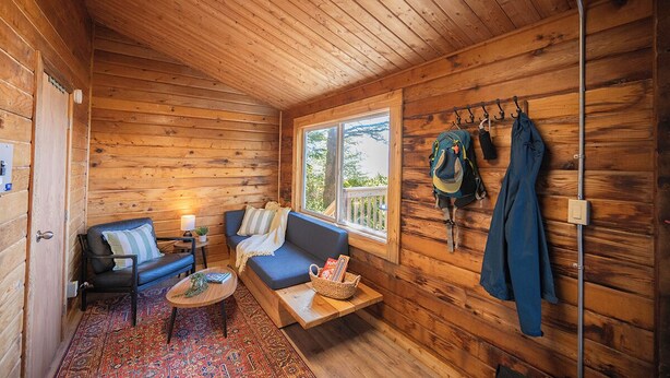 Interior acolhedor de uma cabana com um sofá, uma mesa pequena, uma janela com vista e um cabideiro com itens pendurados.