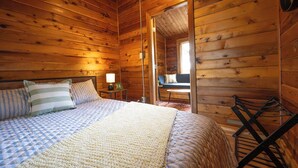Quarto de cabana de madeira com cama, almofadas, mesa de cabeceira, abajur e vista para o exterior.