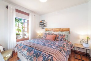 Une chambre avec un grand lit, une table de chevet avec une lampe, une fenêtre avec des rideaux et une plante en pot.