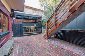 Une terrasse en briques avec un escalier en bois, un espace extérieur couvert aménagé en salon, et un bâtiment à la façade rouge.