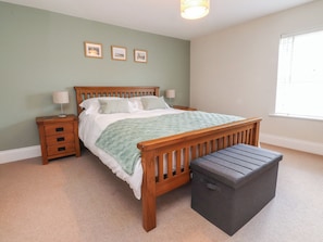A bedroom with a wooden bed, a nightstand, a lamp, a bench, and three framed pictures on the wall. - Grape Lane Cottage (Leyland)