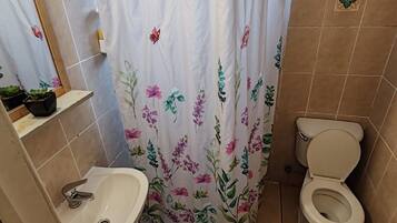 Casa de banho com cortina de chuveiro, pia, sanita e uma pequena prateleira com artigos de higiene pessoal.