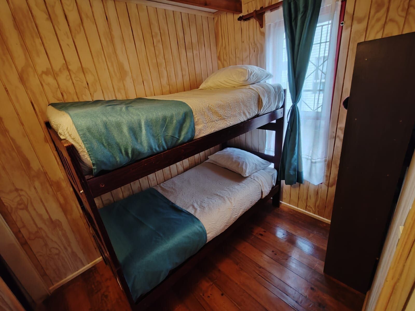 A bunk bed with a green blanket and white bedding in a room with wooden walls and a wooden floor.