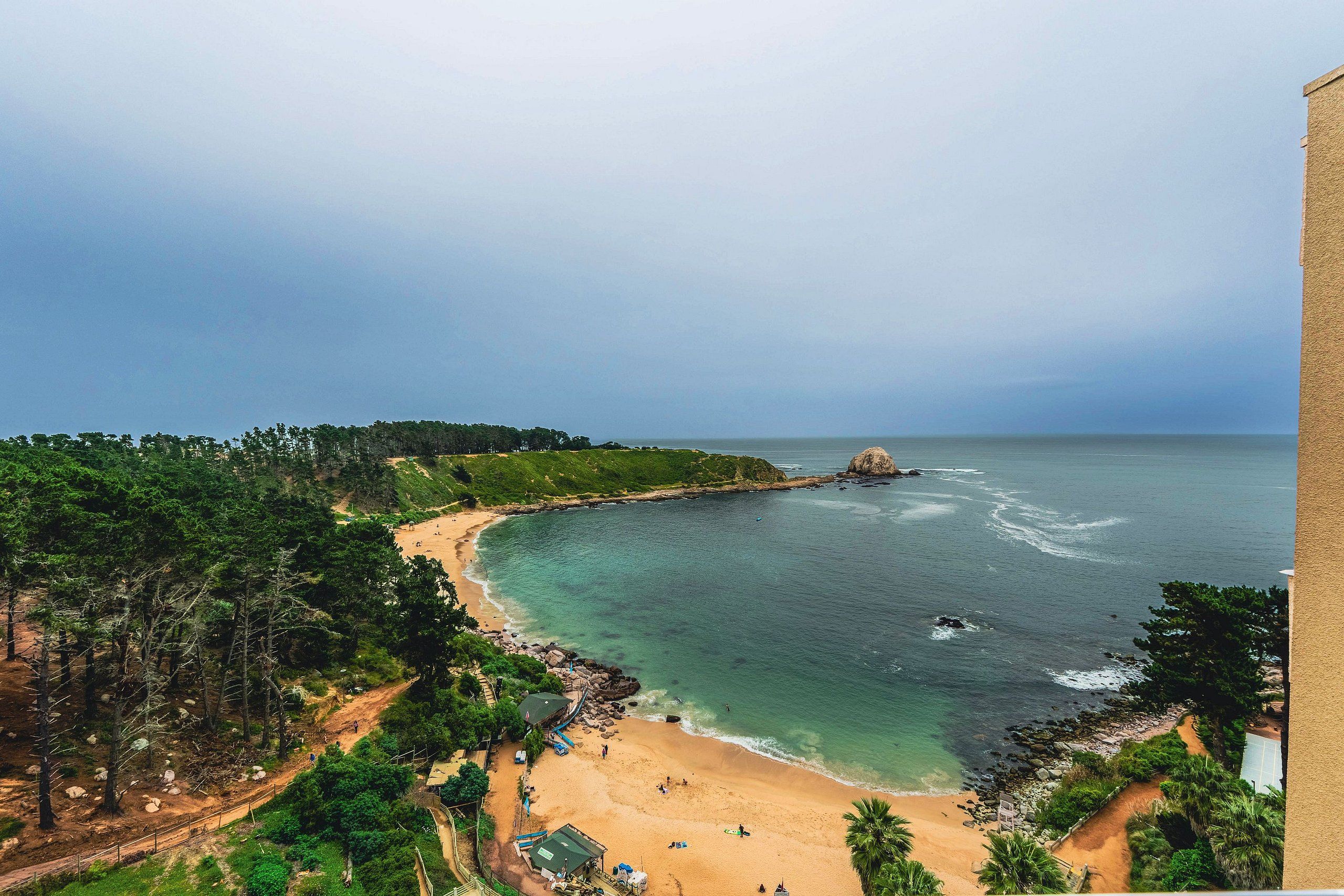 Uma praia costeira com águas cristalinas, um afloramento rochoso e uma área arborizada.