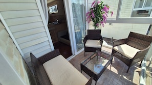 Un patio con mobiliario de mimbre y una mesa de vidrio, una puerta corrediza de vidrio que da a una cocina, y una planta en maceta con flores rosas.