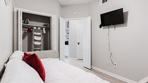 Une chambre à coucher avec un lit, une armoire où sont suspendus des vêtements, un téléviseur fixé au mur et une porte donnant sur une autre pièce.