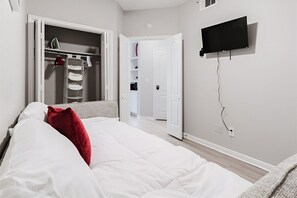 A bedroom with a bed, a closet with hanging clothes, a television mounted on the wall, and a door leading to another room. - SAVE BIG? Walkable Uptown Dallas 2BR/ Nightlife (Dallas)