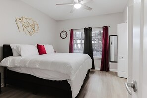 Un dormitorio con una cama, un ventilador de techo, una ventana con cortinas, un reloj y una puerta.