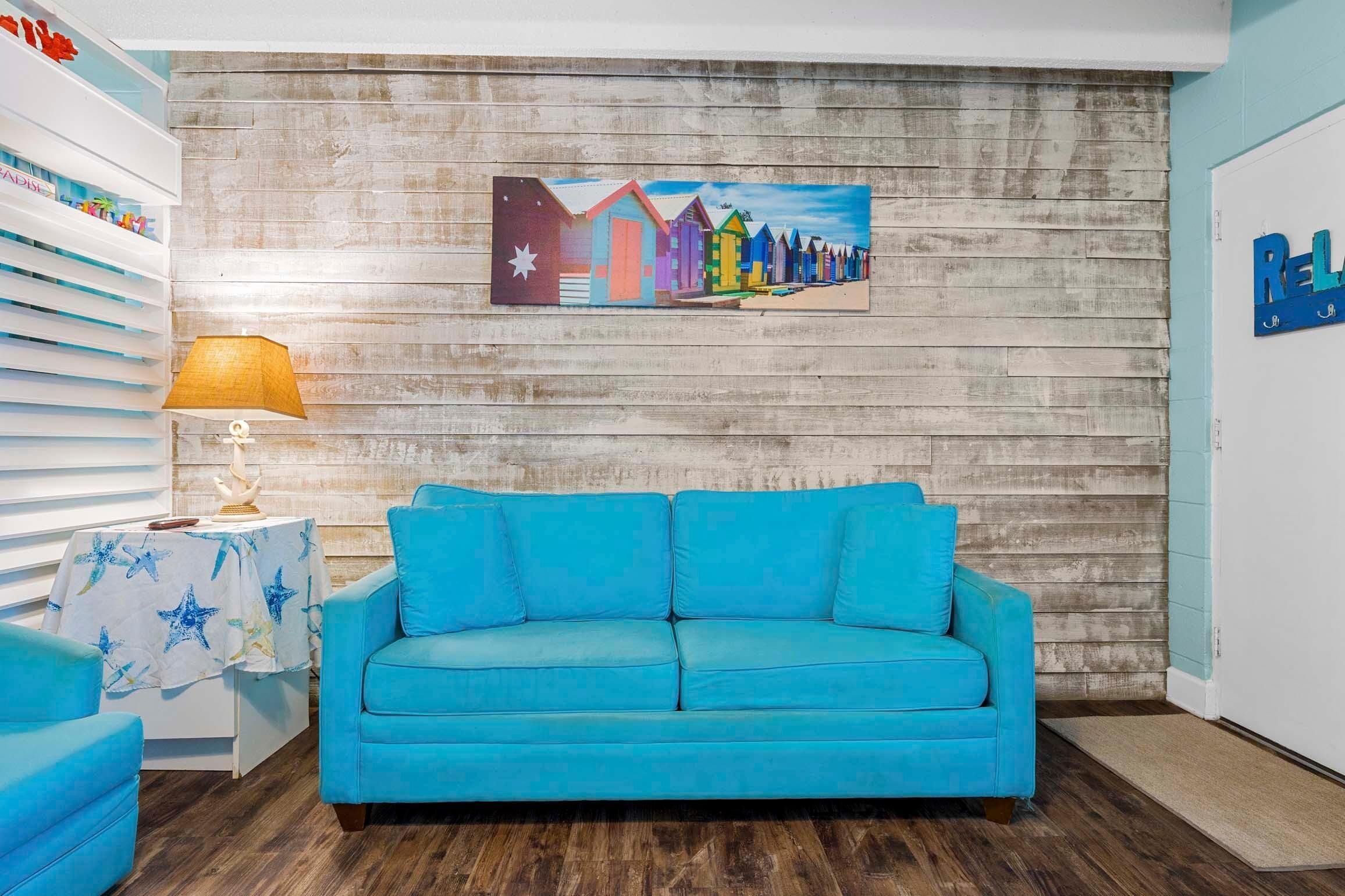 A room with a blue sofa, a wooden wall, a starfish decoration, and a painting of colorful beach huts.