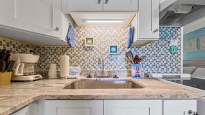 A kitchen with a marble countertop, white cabinets, a stainless steel sink, and a tiled backsplash. - Gulf Village 101 (Gulf Shores)