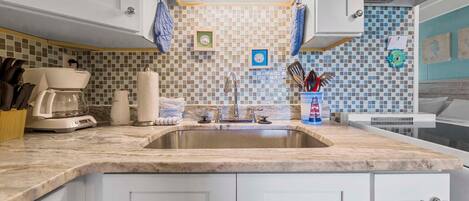 A kitchen with a marble countertop, white cabinets, a stainless steel sink, and a tiled backsplash.