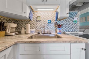A kitchen with a marble countertop, white cabinets, a stainless steel sink, and a tiled backsplash. - Gulf Village 101 (Gulf Shores)