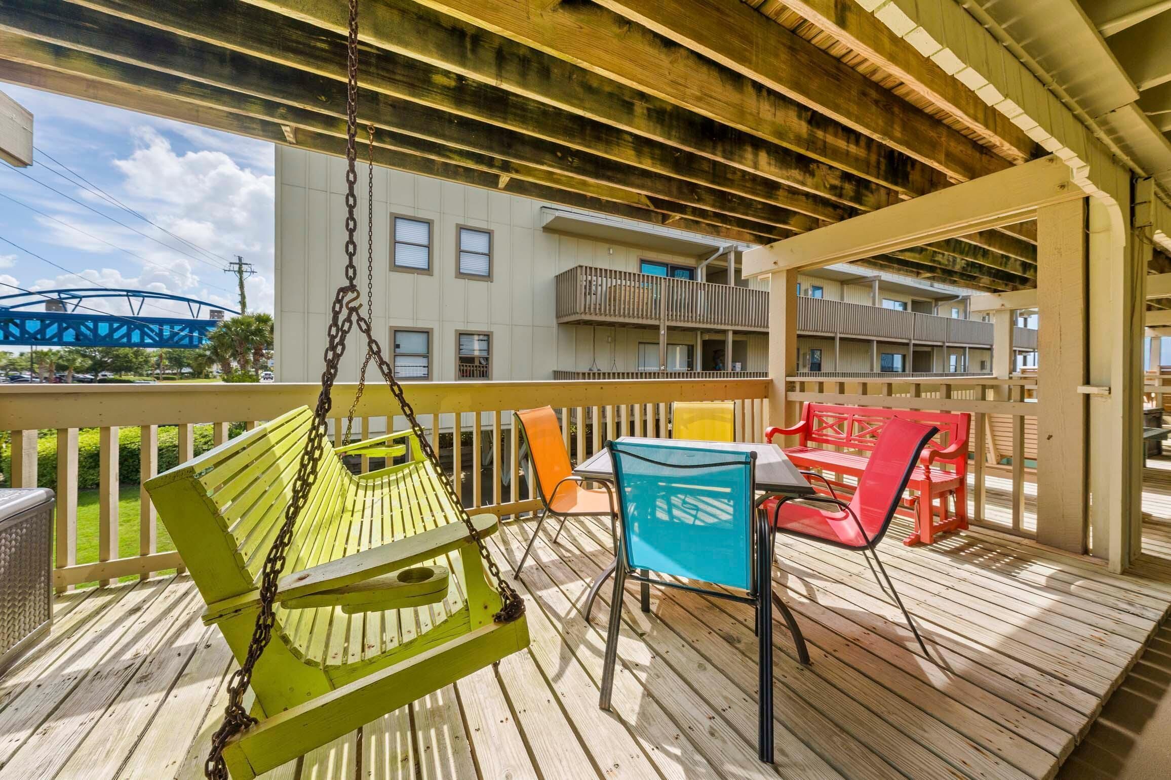 Una terraza de madera con una hamaca, una mesa con sillas y vistas a un edificio con balcones.