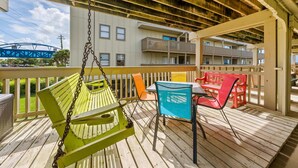 A wooden deck with a swing, a table with chairs, and a view of a building with balconies. - Gulf Village 101 (Gulf Shores)