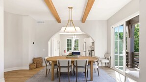 Un espace repas comprenant une table en bois, six chaises, un luminaire suspendu et une vue sur l’extérieur grâce à de grandes fenêtres.