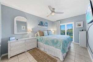 A bedroom with a bed, a dresser, a ceiling fan, a TV, and a window with a view.