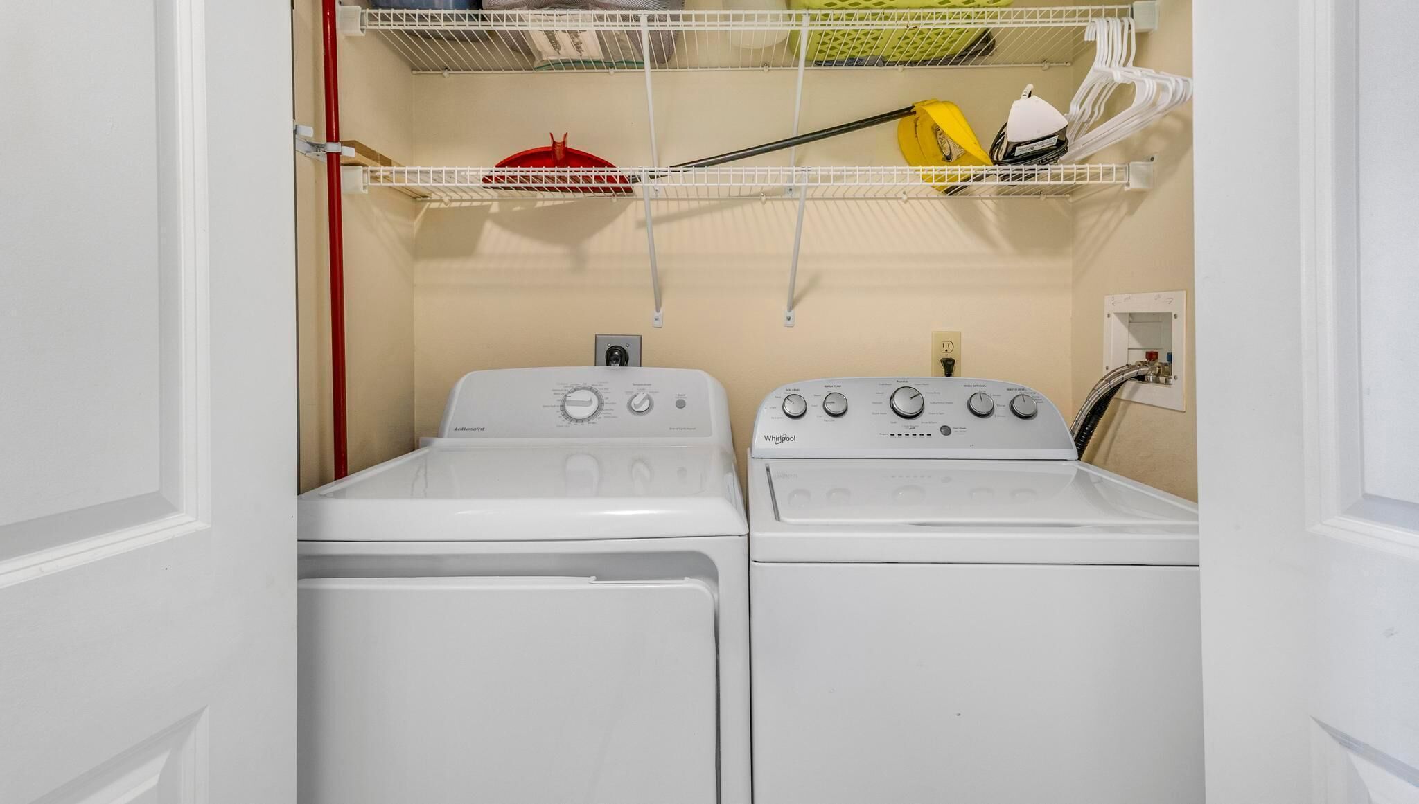 A laundry room with two white front-loading washers and dryers, a shelf with laundry supplies, and a red pipe on the left.