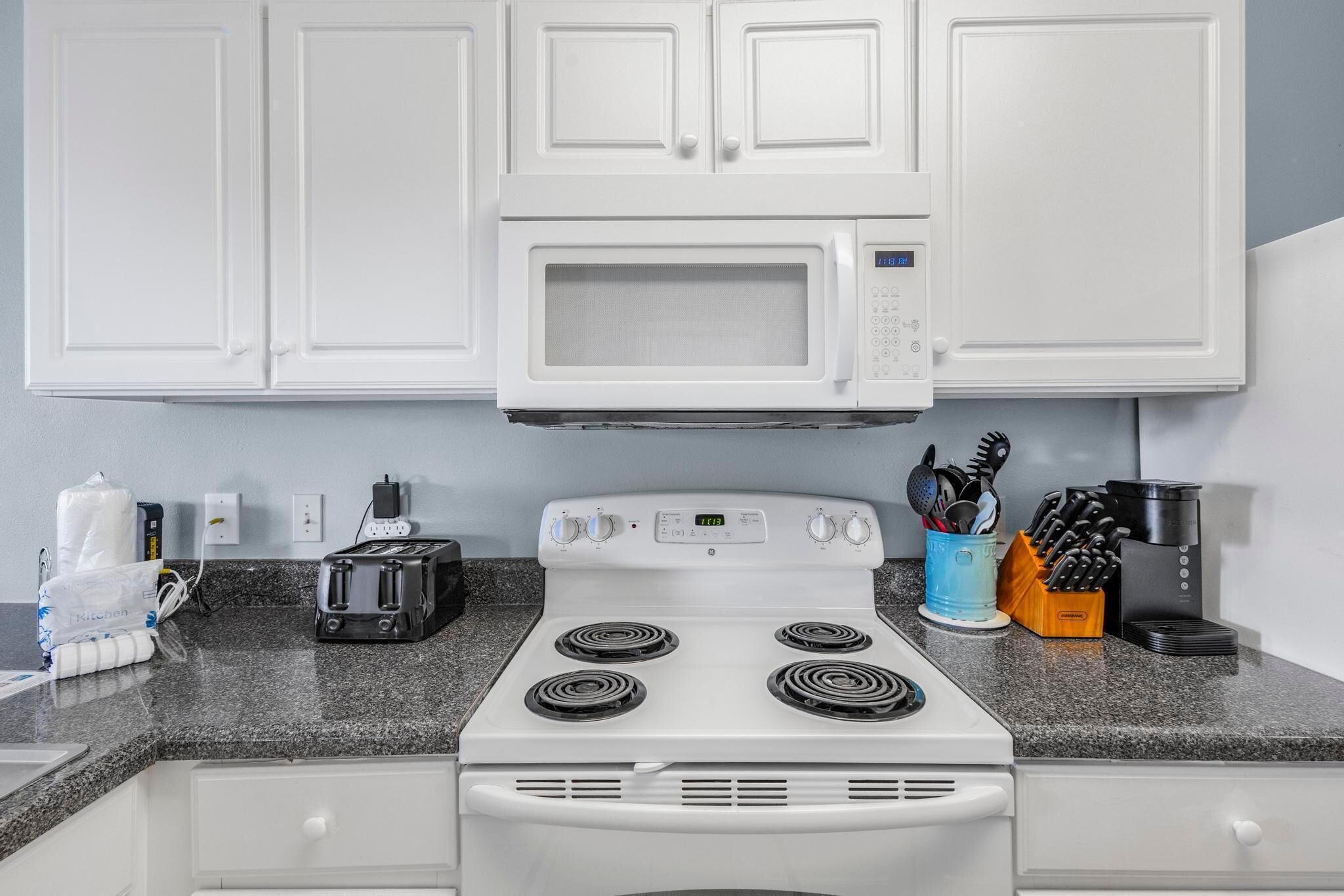 Cocina con armarios blancos, microondas, estufa, tostadora y varios utensilios de cocina.