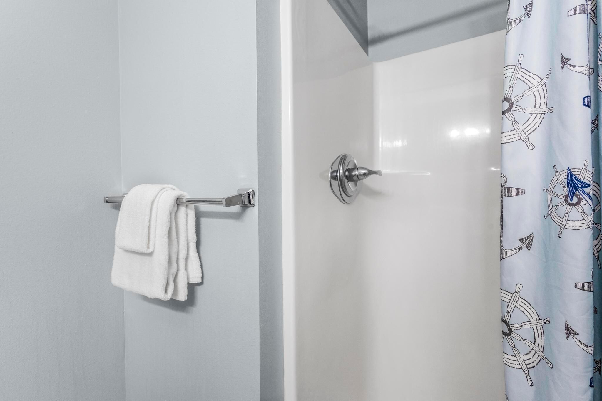Une salle de bain avec une serviette blanche posée sur un porte-serviettes en métal, une porte de douche avec un rideau blanc orné d’un motif nautique et un mur blanc uni.