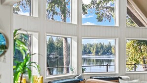 Huoneessa on suuret ikkunat, joista avautuu näkymä järvelle ja metsään. Huonekalut ovat puuta, ja siellä on myös keltainen tuoli.