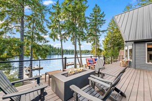 A deck with outdoor seating, a fire pit, and a view of a lake surrounded by trees.