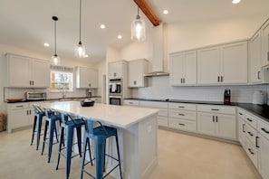 A modern kitchen with white cabinets, a large island, blue bar stools, and pendant lighting. - Secluded Hill Country Getaway • Stunning Views + Fire Pit • 15 Min to Main St (Fredericksburg)