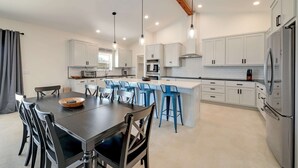Uma cozinha moderna com armários brancos, uma ilha central com banquetas azuis, uma mesa de jantar com cadeiras e uma geladeira grande.