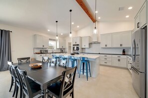 A modern kitchen with white cabinets, a central island with blue bar stools, a dining table with chairs, and a large refrigerator. - Secluded Hill Country Getaway • Stunning Views + Fire Pit • 15 Min to Main St (Fredericksburg)