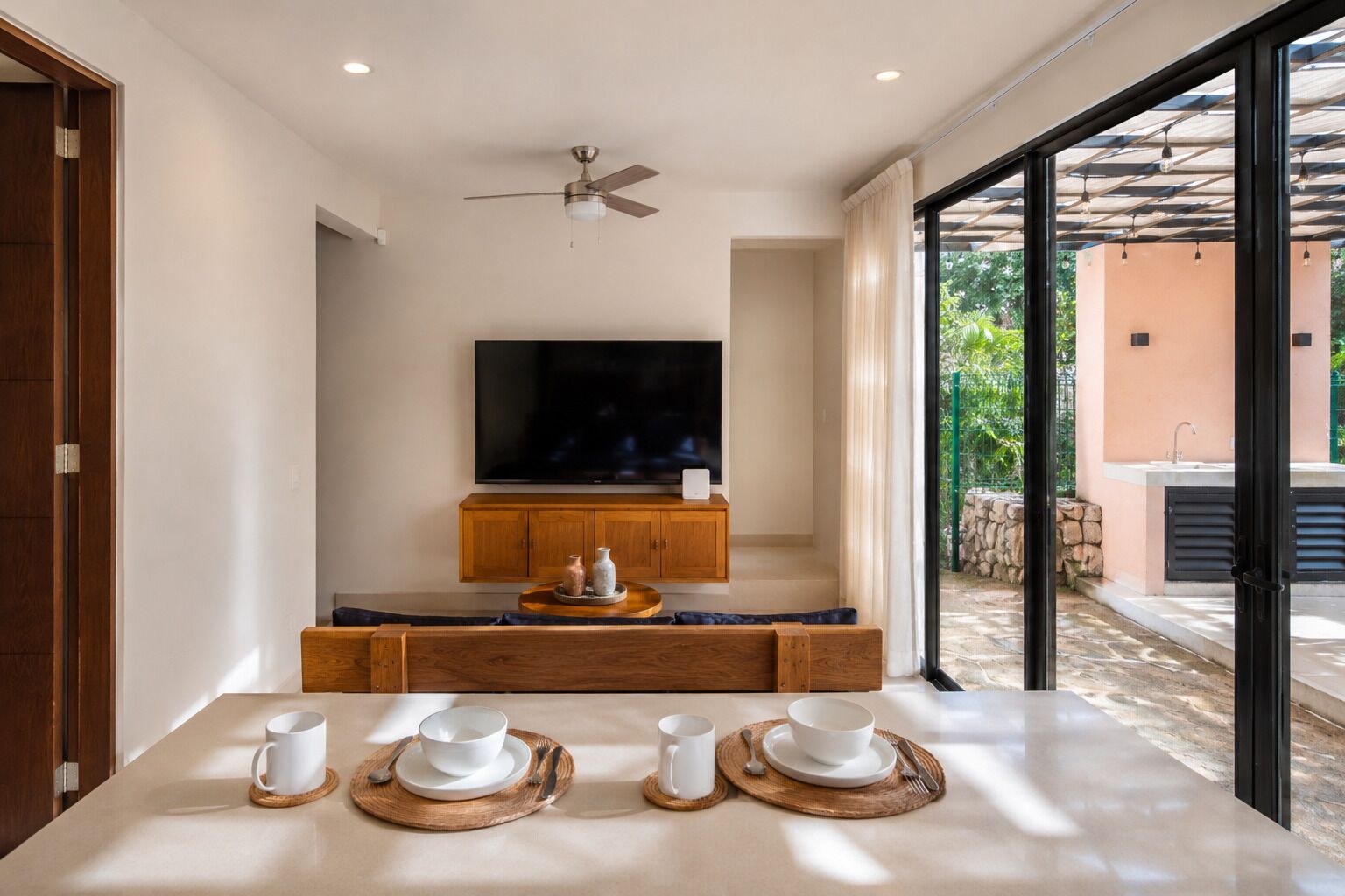 A modern living room with a flat-screen TV, wooden cabinets, and a dining area set with white tableware.