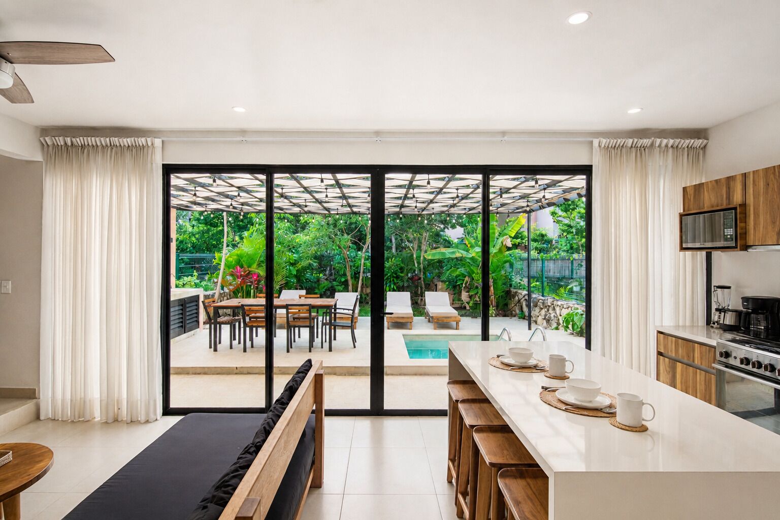 Une cuisine moderne dotée d’un grand îlot central, de portes-fenêtres coulissantes donnant sur un patio, et offrant une vue sur une piscine et un jardin.