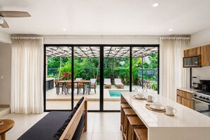 A modern kitchen with a large island, sliding glass doors to a patio, and a view of a pool and garden. - Tropical 3BR Villa Retreat + Private Plunge Pool (Tulum)