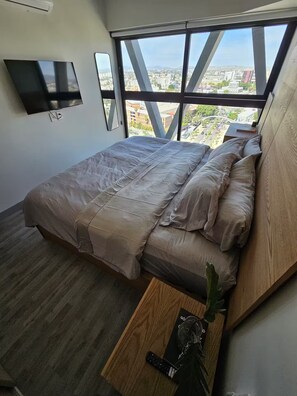 Une chambre moderne avec un grand lit, un téléviseur fixé au mur et une vue sur la ville grâce à de grandes fenêtres.