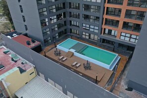 Une piscine sur le toit, dotée d’une terrasse en bois et de chaises longues, entourée d’immeubles à plusieurs étages avec de grandes fenêtres.