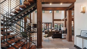 Un intérieur moderne avec un escalier en bois, un espace de vie comprenant un canapé et des fauteuils, et une vue dégagée grâce à de grandes fenêtres.